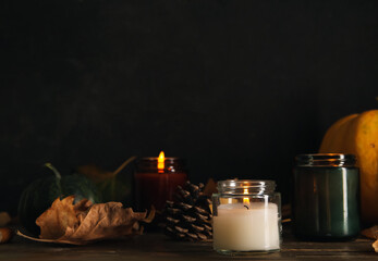 Burning candles with autumn leaves and pine cone on table near black wall