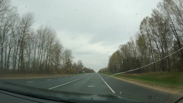 First-person view of a passenger in the windscreen of a car. A crack in the windscreen. It's raining lightly, wipers are working. Grey sky and bare trees. Early spring. Travelling by car