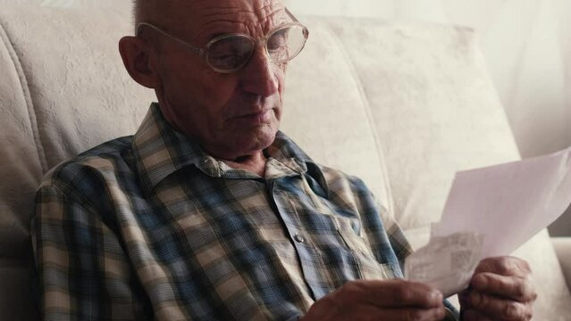 Old senior man looks with surprise on utility bills. Planning month budget, calculating expen. Wrinkled palm of close up.