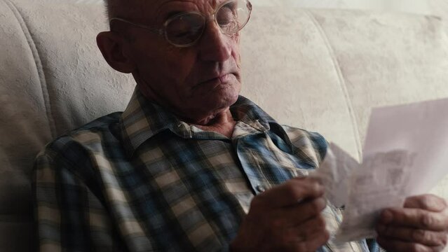 Old senior man looks with surprise on utility bills. Planning month budget, calculating expen. Wrinkled palm of close up.
