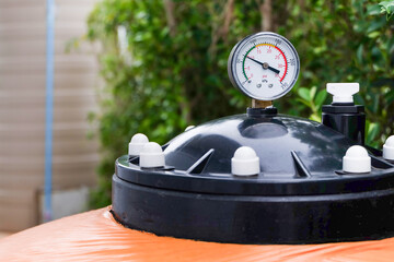 Outdoor swimming pool filtration system, pressure gauge measuring water pressure on top of an orange color sand filter tank
