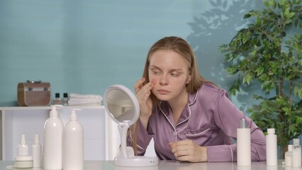 A young woman without makeup, sitting at her dressing table in the morning, looks at her face in...