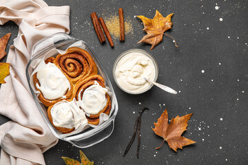Baking dish of tasty cinnamon rolls with cream and autumn leaves on black background