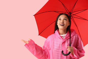 Beautiful young Asian woman in raincoat with umbrella pointing at something on pink background