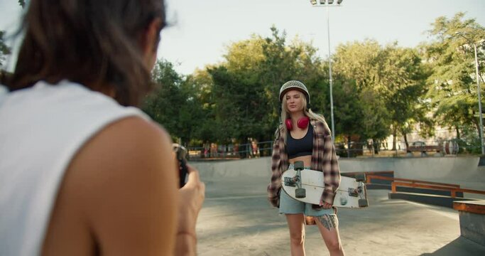 A brunette guy in a white T-shirt takes a photo of a blonde girl in a checkered shirt with a skateboard on a camera that immediately prints a photo in a skatepark in the summer