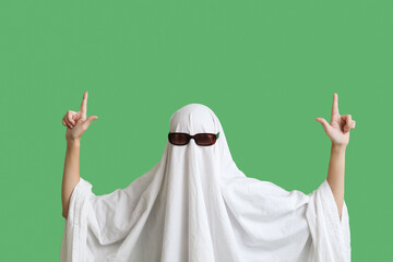 Woman in Halloween costume of ghost and sunglasses pointing at something on green background