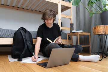 A teenager is doing homework sitting on the floor with a laptop and a book.