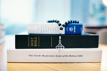 Books, stack and closeup on table for faith, Abrahamic religion or rosary with crucifix for study in home. Knowledge, holy spirit and education with cross for Jesus, Muhammad or Moses with solidarity - obrazy, fototapety, plakaty