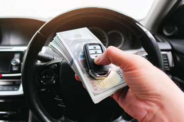 Driver hand holding a car key or keyless remote and usd dollar banknotes with a steering wheel on background in a car. Buy sell and car loan concept - obrazy, fototapety, plakaty