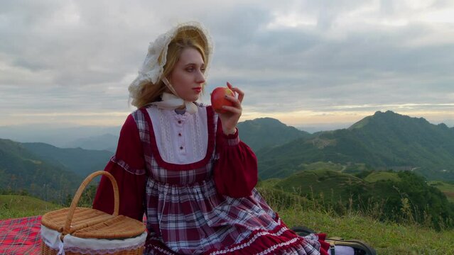 Medium view of woman picking up apple from red picnic blanket overlooking mountain vista