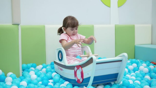 Excited Girl Toddler Sitting in Toy Boat And Tries to Catch Balls From Dry Pool Which Mom Throw in Kids Playroom