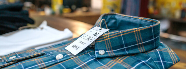 Detail of label with price and size over a blue plaid shirt on industrial style store. Banner of...