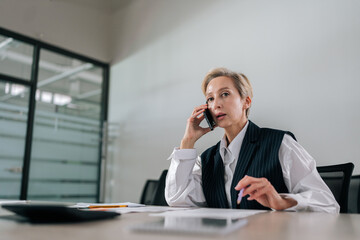 Low-angle view of middle-aged businesswoman making business call sitting on office desk. Portrait of gray-haired female manager talking at smartphone with client discussing contract.