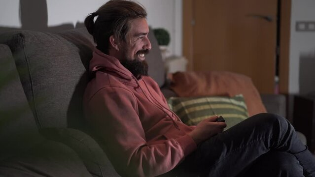 Side view cheerful bearded Caucasian man sitting happy on sofa laughing. Young casual millennial generation male smile enjoying funny comedy movie or series and turning on TV indoors at night. 