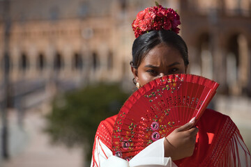 Portrait of a young black and South American woman in a beige flamenco gypsy costume and red shawl, covering herself from the sun with a fan in Seville in Spain. Concept dance, folklore, flamenco, art