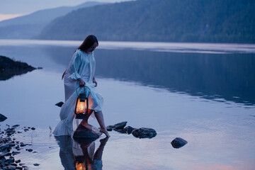 Young Asian girl in nightgown with retro lamp on stones on river