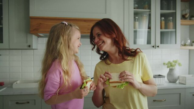 Positive mother with daughter stand in the kitchen with sandwiches they have prepared and demonstrate tenderness to each other. Mom and child pose with a smiles and look at the camera. The concept of