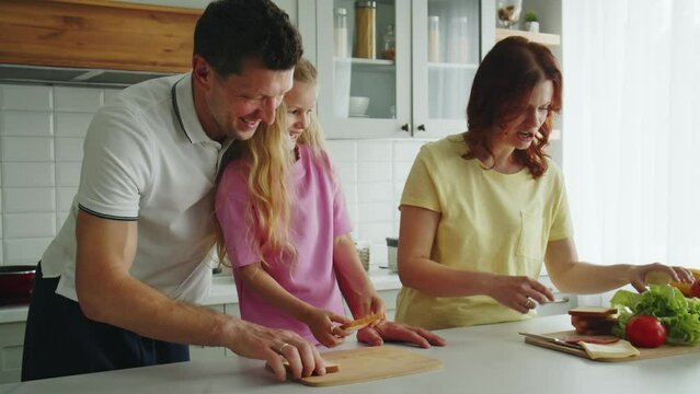 A positive smiling american dream family in the kitchen make sandwiches playfully. The daughter helps mom and dad to prepare a snacks for a picnic or guests. The healthy eating demonstration. 