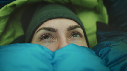Woman wake up in camp tent sleeping bag. Close up portrait of tourist girl opening eyes in cold morning. Female traveler wilderness vacation, tourism and camping in outdoor trip. - obrazy, fototapety, plakaty