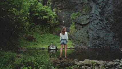 Obraz premium Tourist woman enjoy jungle waterfall landscape standing on rock stone. Girl relax outdoor lifestyle travel on summer holiday vacation. Picturesque scene panoramic shot. Dramatic dark toning, back view
