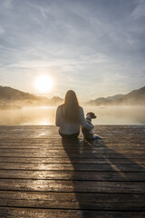 Rückansicht von einer jungen Frau und einem kleinen Terrier Hund auf einem Holzsteg an einem See. Sonnenaufgang, Nebel.