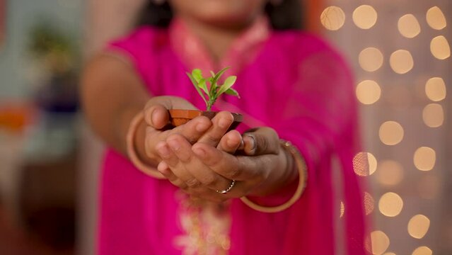 close up shot of Indian young girl kid showing diya with plant by looking camera during diwali celebration - concept of environmental protection, save plants and stop festive pollution
