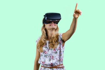 Happy surprised mature woman in virtual reality headset pointing up. Isolated on turquoise.