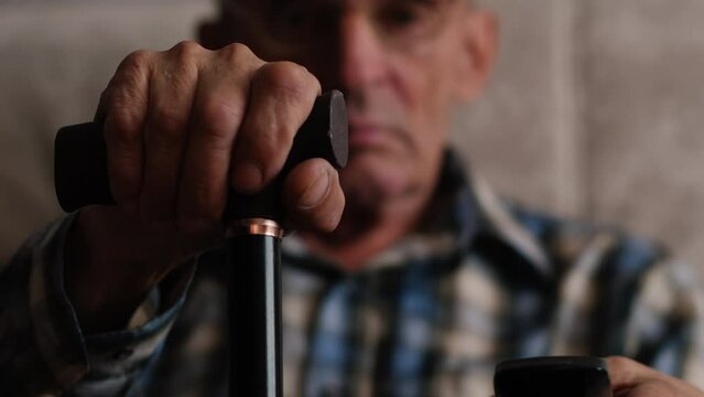 Close up side profile photo of grey hair he his him grandpa leaning with grief on walking stick looking straight oldness in mind casual checkered shirt sitting on cozy divan