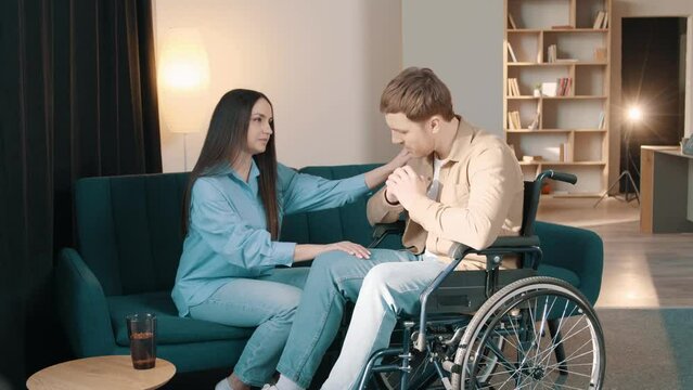 caring young wife putting hand on husband sitting in wheelchair at home, young married couple support each other