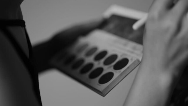 Closeup grayscale shot of the hands of a makeup artist holding an eyeshadow palette and using it