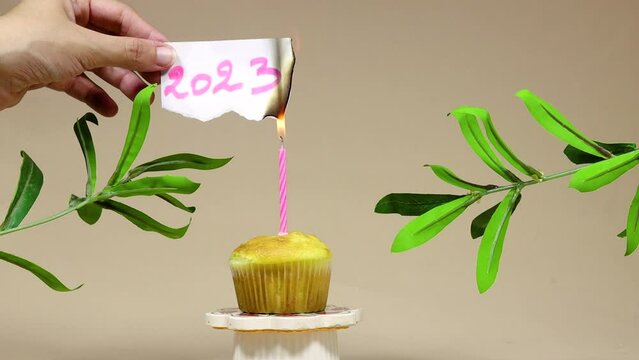 closeup hand burning a paper with 2023 text meaning goodbye and welcome a new year 2024, candle of birthday cupcake or madeleine above podium with ruscus plants