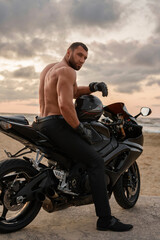 Handsome ripped man sits on his black sports motorcycle at a desolate beach, against a cloud-laden sunset
