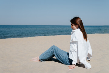 Fototapeta na wymiar Side view of a lonely pensive young woman in casual clothes sitting on sand on seashore