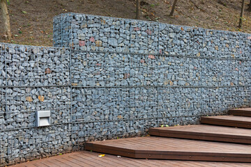 Gabion fence with gravel in a metal structure. Modern stone wall and wooden steps of a staircase in...