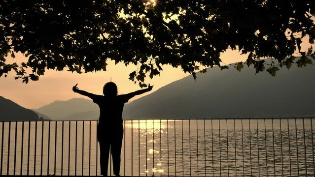 Woman in Silhouette Walking on the Waterfront with Arms Raised and Railing and Tree Branch over Lake Lugano with Mountain in Sunset in Bissone, Ticino, Switzerland.