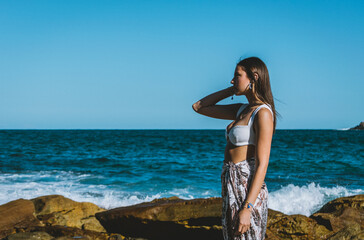 young woman modeling on the beach on the rocks with the sea in the background. dressed in a sarong and bikini with her hair flying. standing showing the jewelry she wears on her legs, arms, and earrin