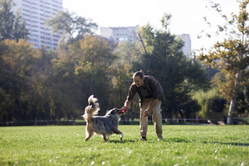 A young white-skinned man with long hair plays with his shepherd dog in the park