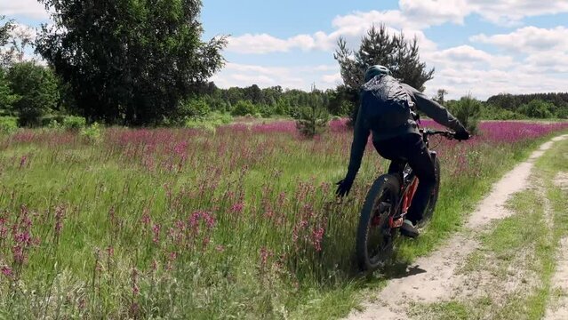 Backpacker sport man extreme driving on bike at meadow path touching blossom spring summer flowers. Follow shot male mountain bicycle speed riding natural sun light day field with floral grass 4k