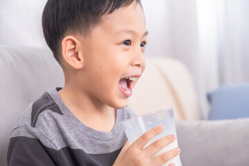 Close-up Asian preschool boy with milk mustache smile at unrecognizable mother sitting on sofa at home, young kid happy hold and drinking a clear glass full of dairy milk, healthy children's lifestyle