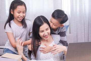 Happy single mom with children playing together bonding relationship at home, Asian freelancer woman working at home using modern technology laptop, kids sibling run to hug mother from behind and kiss