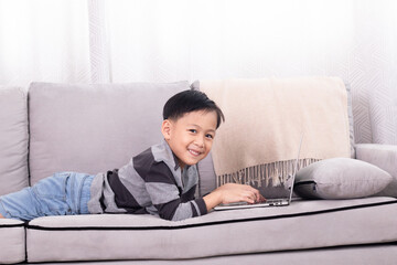 Asian sibling little boy playing game alone at home lying down on sofa using modern technology wireless laptop computer addiction, cute son studying homeschool typing keyboard watching digital lesson
