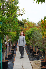 A young woman stands in a large greenhouse and chooses a pot with a small tree. A woman chooses plants for landscaping the yard