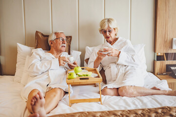 An elderly married couple is getting comfortable in the hotel room, drinking coffee in bed and wearing bathrobes