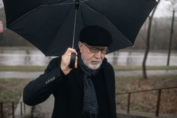 An elderly man in a black coat with an umbrella stands on the stairs in the rain on the background of the road, rainy weather