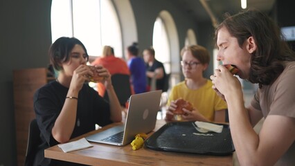 group of students sit in a cafe and do a project on a laptop together. business concept of modern training and lifestyle development. students discuss their homework and eat fast food burgers