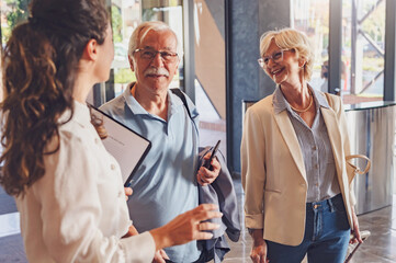 A senior couple is arriving at the hotel with luggage, and a smiling hotel representative is...
