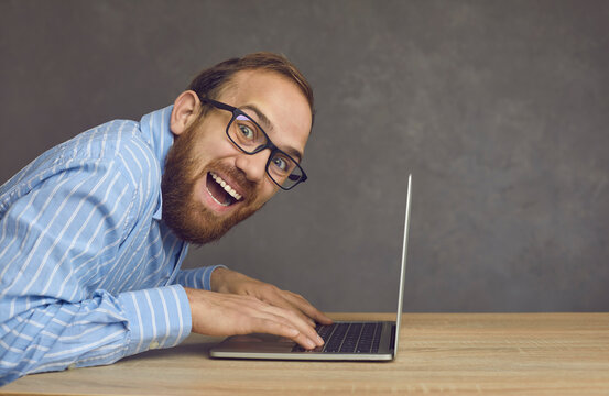Side view portrait of a joyful crazy hacker or programmer sitting at a table typing on a laptop. Funny bearded adult man with glasses on a gray background. Business, cybercrime and web design concept.