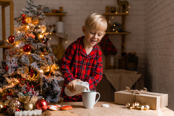 Blond boy in red plaid pajamas drinks milk in the Christmas kitchen. Healthy breakfast