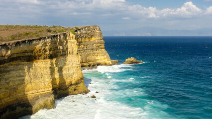 Sandy cliff with grass over blue ocean with white waves, rocks, blue sky, clouds and mountains...