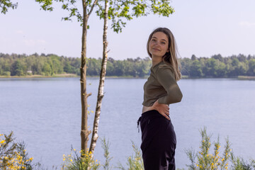 A beautiful and charming young millennial brunette stands on the serene shores of a forest lake....